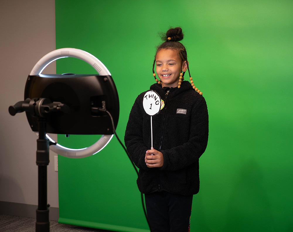 A girl holds a prop in front of a green screen as a camera takes her picture